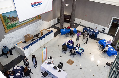 As seen from above people are gathered seated and standing at folding tables arranged for a job fair in a lobby with a marble floor.